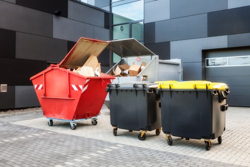 In the foreground, three large wheeled waste bins are positioned on a paved surface adjacent to a modern building with dark grey cladding; the first bin is red with a partially open lid, revealing cardboard boxes and paper waste inside, while the other two bins are black with closed lids. The red bin appears to be made of durable plastic with a textured surface and red-and-white reflective markings on its sides. The black bins are similarly constructed, with smooth surfaces and bright yellow lids on one of them. Behind the bins, a grey metal container with an open lid is visible, also filled with cardboard and paper waste. The environment suggests an urban setting, possibly near a commercial or office building in Rotherhithe, with clean paving and contemporary architectural features. This scene relates to waste management services offered by Business Waste Removal Rotherhithe, demonstrating typical rubbish collection and disposal methods for business premises in the area.