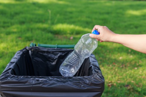 A person’s hand is seen placing a transparent plastic water bottle with a blue cap into a black bin lined with a black plastic bag, positioned outdoors on a grassy area. The background features a well-maintained lawn with bright green grass, indicating a suburban or park setting. The plastic bottle appears to be empty, with some light reflections on its surface, and the person's hand is visible from the wrist to the fingertips, with a natural skin tone. The scene depicts typical waste disposal activity suitable for a rubbish removal service in Rotherhithe, emphasizing proper waste segregation and recycling considerations. The environment is brightly lit with natural sunlight, highlighting the clarity of the plastic bottle and the texture of the bin’s lining material. This imagery aligns with professional rubbish collection and waste management services, illustrating an everyday waste disposal task in a clean, outdoor environment.