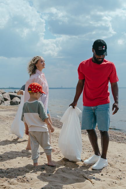A man in a red T-shirt, light blue denim shorts, and a navy cap is standing on a sandy beach, holding a white rubbish bag in each hand while engaging in a beach clean-up activity. Nearby, a woman with