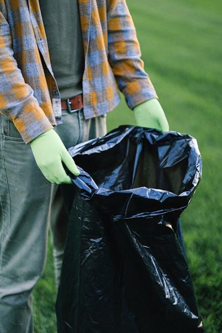 A man in a red T-shirt, light blue denim shorts, and a navy cap is standing on a sandy beach, holding a white rubbish bag in each hand while engaging in a beach clean-up activity. Nearby, a woman with