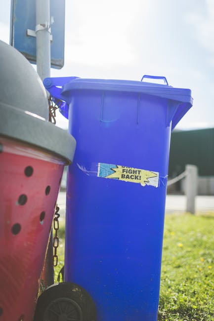 A middle-aged man with dark hair wearing a black T-shirt and dark trousers is seen discarding waste into a cylindrical public rubbish bin made of shiny stainless steel with a slightly domed lid, situa