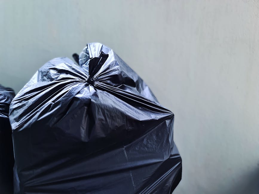 A close-up view of several large black plastic rubbish bags filled with waste, piled together against a plain, light-colored wall. The bags have a glossy, crinkled texture and are tightly closed with 