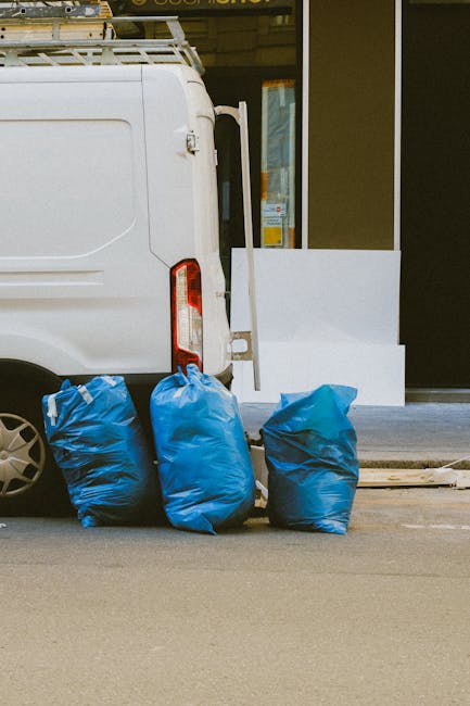 A close-up view of several large black plastic rubbish bags filled with waste, piled together against a plain, light-colored wall. The bags have a glossy, crinkled texture and are tightly closed with 