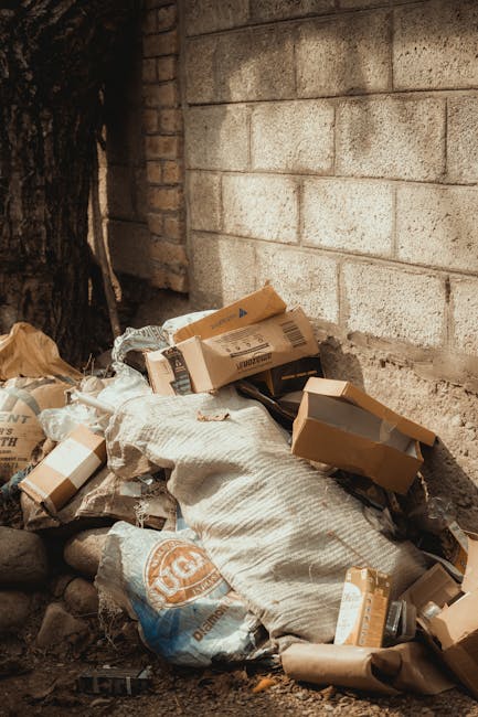 A person wearing a light blue T-shirt, black shorts, and white sneakers is using a grabber tool to pick up small pieces of rubbish or debris from a rocky outdoor area. The rocks are varied in size, pr